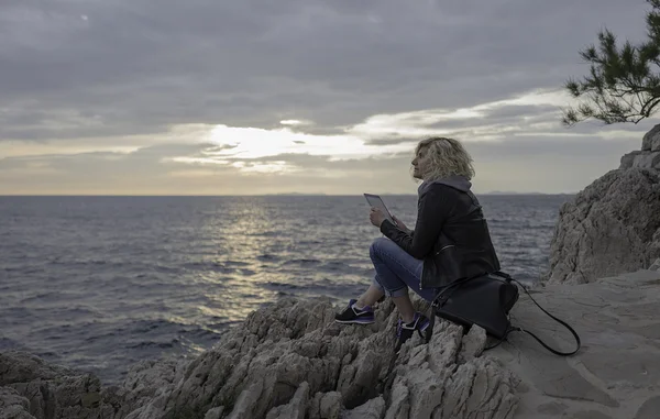 Krásná blonďatá dívka je sezení na kameny u moře s tablet v ruce a díval se do dálky. — Stock fotografie