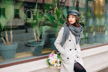 Beautiful woman sits on the cornice by the window with a bouquet of color and looks into the distance