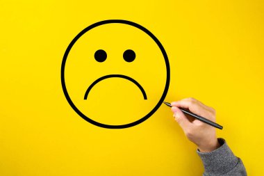 Erkek el, sarı arka planda üzgün bir yüz çizimi yapıyor. Müşteri memnuniyeti, hizmet veya ürün değerlendirme kavramı.