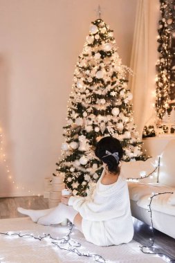 Beyaz kazaklı bir kadın, Noel iç tasarımında sıcak ve rahat bir akşam, ışıklarla süslenmiş Xmas ağacı hediyeler, mumlar, kapalı mekanda çelenkler. Tatil salonu. Yeni Yıl
