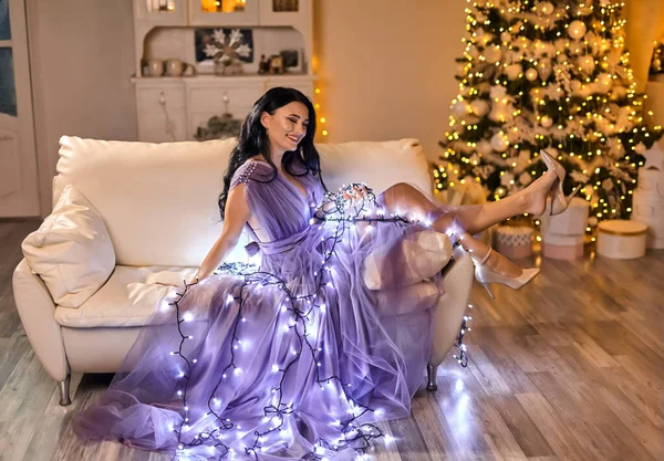 beautiful fairy woman wearing elegant fashionable evening dress posing on sofa in warm magic evening in luxury Christmas living room, decorated by garland light