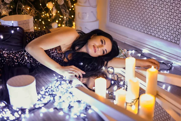beautiful fairy woman wearing elegant fashionable evening dress posing on the floor in warm magic evening in luxury Christmas living room, decorated by garland light and candles