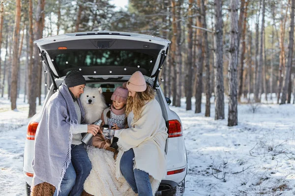 Happy family with dog on vacation during the winter holidays near road. Dressed in warm clothes sitting on the trunk of a car and drinking tea from a thermos. space for text. Winter vacation