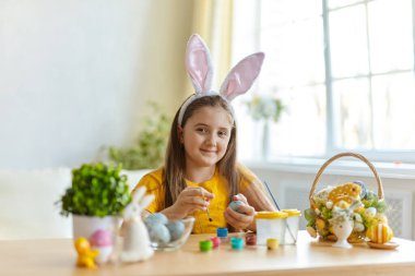 Tavşan kulağı takan şirin bir çocuk. Oturma odasındaki masada oturan kız. Çocuk yumurta boyuyor..