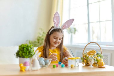 Tavşan kulağı takan şirin bir çocuk. Oturma odasındaki masada oturan kız. Çocuk yumurta boyuyor..