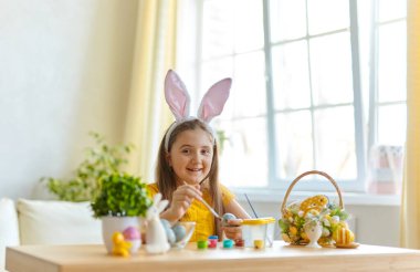 Tavşan kulağı takan şirin bir çocuk. Oturma odasındaki masada oturan kız. Çocuk yumurta boyuyor..
