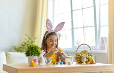Tavşan kulağı takan şirin bir çocuk. Oturma odasındaki masada oturan kız. Çocuk yumurta boyuyor..