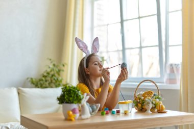 Tavşan kulağı takan şirin bir çocuk. Oturma odasındaki masada oturan kız. Çocuk yumurta boyuyor..