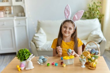 Tavşan kulağı takan şirin bir çocuk. Oturma odasındaki masada oturan kız. Çocuk yumurta boyuyor..