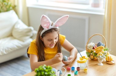 Tavşan kulağı takan şirin bir çocuk. Oturma odasındaki masada oturan kız. Çocuk yumurta boyuyor..