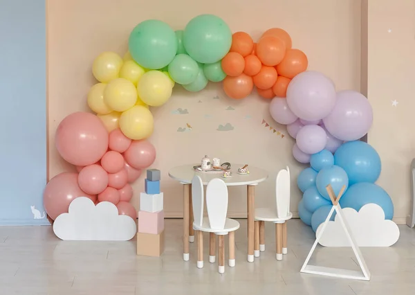 Small table and chairs with bunny ears in children\'s room interior. Rainbow, colorful balloon arch, decoration in honor of the holiday, birthday, party