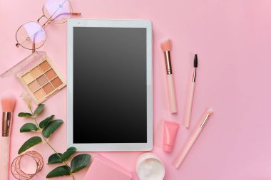 Düz, üst manzara, modern ofis masası. Beyaz tabletli çalışma alanı, not defteri, gözlükler, kalemler, güzellik aksesuarları, klavye, ofis malzemeleri, pembe arka planda yeşil yaprak