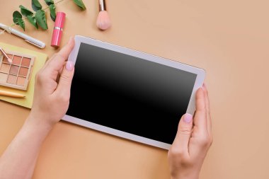 Blogger 's Flalat lay, masa masası. Kadının ellerinde boş tablet, güzellik aksesuarları, bej arka planda yeşil yaprak.