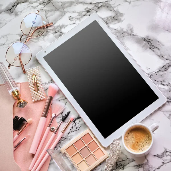 Flat lay composition with tablet and makeup products, coffee and eyeglasses for woman on marble desk