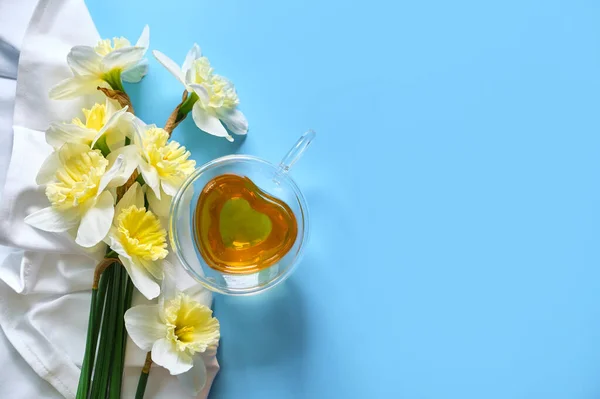 ハート型のグラスカップの中の緑茶で ナプキンの底が2倍になり 青い背景にダフォディの花が付いています 春の静物 — ストック写真