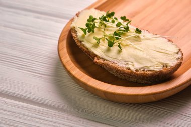 Tahta bir tabakta tereyağlı ve mikroyeşillikli ekmek dilimi, açık ahşap bir arka planda.