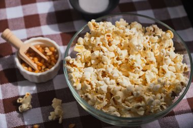 Ev yapımı patlamış mısır yapıyorum. Kareli masa örtüsünün üzerinde cam bir kasede lezzetli bir atıştırmalık. Film izlemek için atıştırmalık.