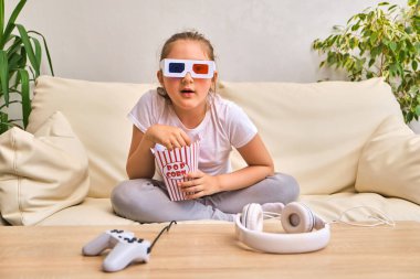 çocukluk ve ev konsepti - üç boyutlu film gözlüklü duygusal küçük kız çizgili kovadan patlamış mısır yiyor ve evde televizyon izliyor.