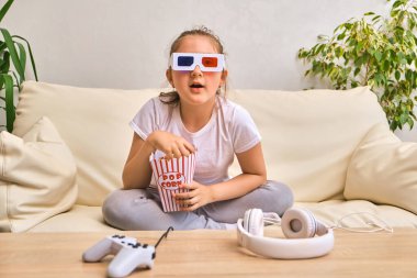 Çocukluk ve ev kavramı. Üç boyutlu film gözlüklü küçük kız çizgili kovadan patlamış mısır yiyor ve evde televizyon izliyor.