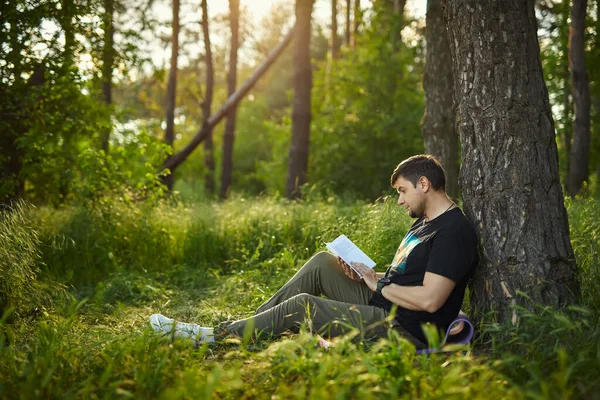 Przystojny Młodzieniec Siedzący Lesie Trawie Opierający Się Drzewo Czytający Książkę — Zdjęcie stockowe