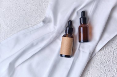 Kehribar rengi cam, beyaz desenli arka planda beyaz kumaş üzerine kahverengi yağ şişeleri. Düz yerde, üst tarafta. Doğal organik güzellik ürünü ambalaj modelleri.