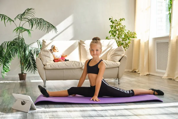 Kid girl exercising at home in a living room. Video lesson. Kid repeating exercises while watching online yoga gymnastics session. Beautiful girl doing leg-split exercise at home