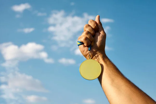 stock image Winner hand raised and holding gold medal with ribbon against blue sky. Golden medal is medal awarded for highest achievement for sport or business. Success Awards concept