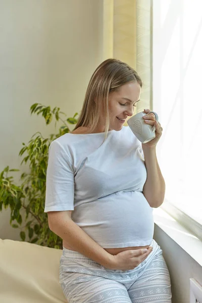 怀孕的年轻女子站在窗边 端着一杯咖啡 — 图库照片