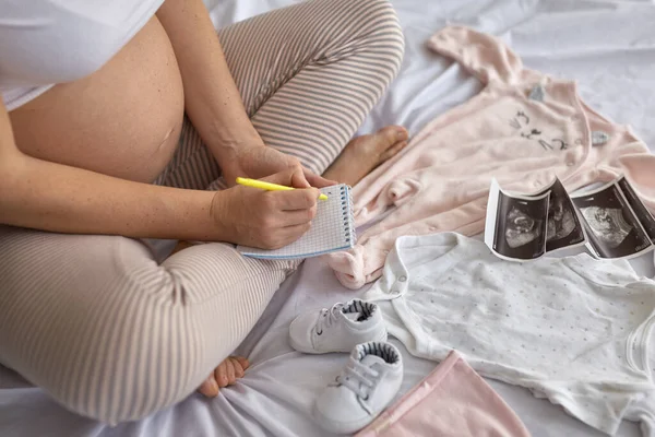 孕妇在床上准备婴儿衣服 在笔记本上填写待办事项清单 怀孕和分娩概念的头像 — 图库照片