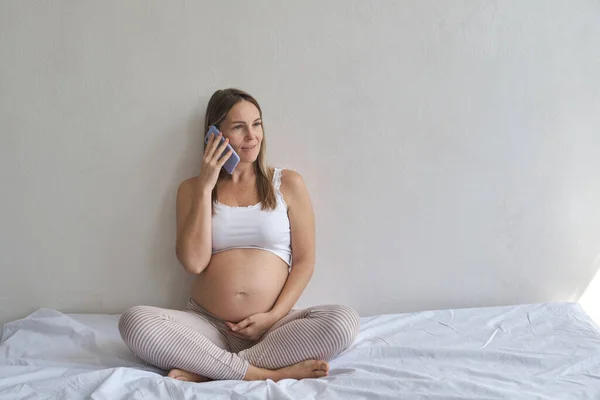 坐在床上 在家中使用智能手机的微笑孕妇 — 图库照片