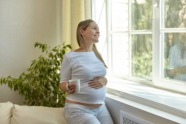 Pregnant young woman stands near the window at home with a cup of coffee