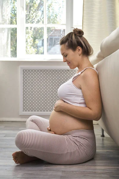 怀孕的女人在家里爱抚她的肚子 年轻的金发孕妇感觉到她的宝宝在推挤 坐在地板上 复制空间 期望概念 — 图库照片