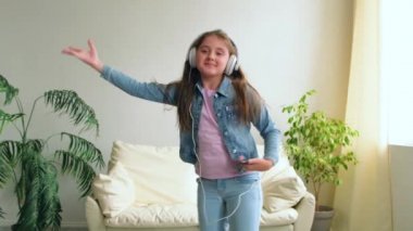 Kulaklıklı komik mutlu küçük kız evdeki koltukta zıplıyor. Kızım oturma odasındaki kanepede akıllı telefonuyla oynuyor. Yaşam Tarzı Evi 'ni seviyorum. Neşeli Komik Çocuk ağır çekimde.