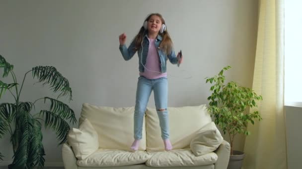 Αστείο Χαρούμενο Κοριτσάκι Ακουστικά Πηδάει Στον Καναπέ Στο Σπίτι Κόρη — Αρχείο Βίντεο