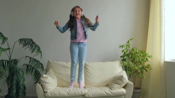 戴着耳机的滑稽快乐小女孩在家里的沙发上跳着 女儿在客厅的沙发上玩智能手机 爱的生活方式的家 慢动作中快乐有趣的孩子 — 图库视频影像