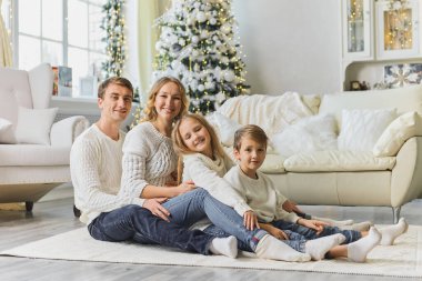 Mutlu Noeller ve mutlu yıllar. Beyaz kazaklı mutlu bir aile oturma odasında yerde birbiri ardına oturuyor ve evdeki güzel Noel ağacının yanında gülüyor. Mutlu bir aile ve iki çocuk.