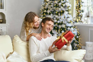 Güzel bir çift birlikte yeni yılı bekliyor, ellerinde hediye kutusuyla, güzel bir Noel ağacının yanında, oturma odasındaki kanepede aydınlatma çelengi ile