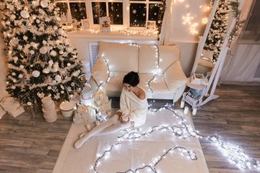 Beyaz kazaklı ve sıcak taytlı bir kadın. Noel iç tasarımında sıcak ve rahat bir akşam. Işıklarla süslenmiş Xmas ağacı. Tatil salonu. Yeni Yıl