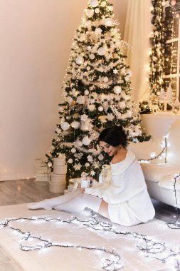 Beyaz kazaklı ve sıcak taytlı bir kadın. Noel iç tasarımında sıcak ve rahat bir akşam. Işıklarla süslenmiş Xmas ağacı. Tatil salonu. Yeni Yıl