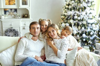 Güzel, mutlu bir ailenin portresi. Beyaz süveterli iki çocuk kanepeye oturmuş Noel ağacıyla süslenmiş bir odada.