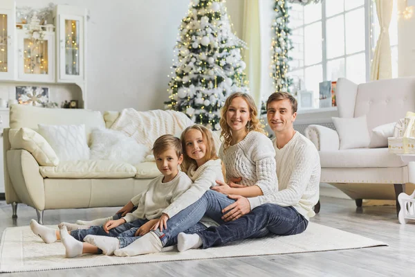 Mutlu Noeller ve mutlu yıllar. Beyaz kazaklı mutlu bir aile oturma odasında yerde birbiri ardına oturuyor ve evdeki güzel Noel ağacının yanında gülüyor. Mutlu bir aile ve iki çocuk.
