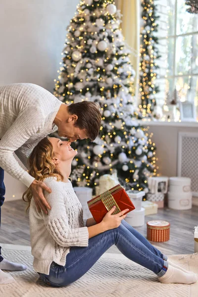 美しいカップルは一緒にお正月を待っています手にギフトボックスで抱き合っている間 男は照明ガーランドと居心地の良いリビングルームのソファで美しいクリスマスツリーの近くに妻にキスをします — ストック写真