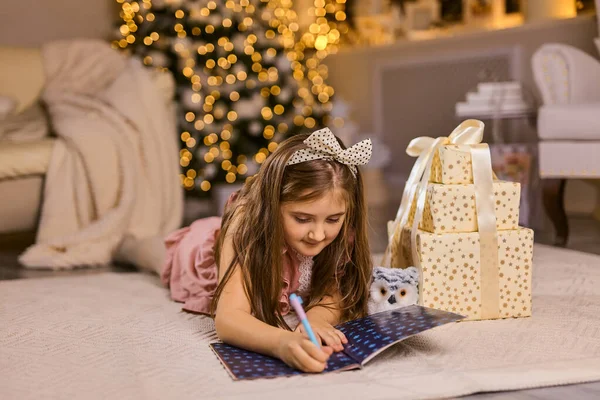 メリークリスマスとハッピーホリデー かわいい子供の女の子は屋内でクリスマスツリーの近くにサンタクロースに手紙を書きます — ストック写真