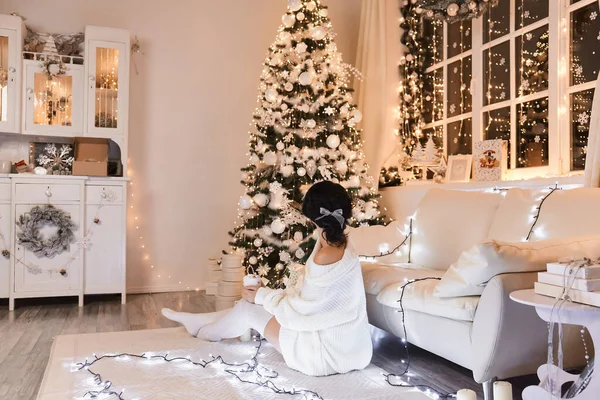 Beyaz kazaklı ve sıcak taytlı bir kadın. Noel iç tasarımında sıcak ve rahat bir akşam. Işıklarla süslenmiş Xmas ağacı. Tatil salonu. Yeni Yıl