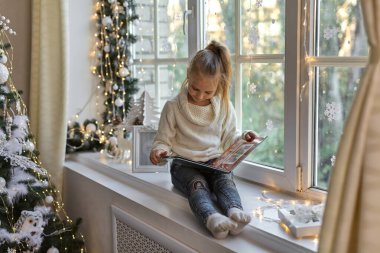Mutlu Noeller ve mutlu Noeller. Pencerenin kenarında oturup kitap okuyan tatlı küçük bir kız. Noel 'de dekore edilmiş bir oda. Çocuk peri masalından hoşlanıyor..
