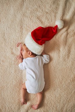 Beyaz tulumlu yeni doğmuş bir bebeğin kabarık bej bir battaniyenin üzerinde göbeğinin üstünde yatışı. Bebek büyük bir Noel Baba şapkası takıyor ve nazikçe elinde kırmızı beyaz bir şeker kamışı tutuyor.