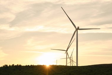 Sıcak turuncu günbatımı gökyüzüne karşı gerçek yaşam rüzgar çiftliği siluetleri, düşük ufuklu çimenli tepelerde türbinler, dramatik altın ışık, kırsal kırsal alanda yenilenebilir enerji manzarası alacakaranlıkta