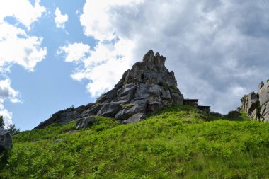 Çimenli bir yamaçta yükselen engebeli kaya oluşumu. Parlak mavi gökyüzünün altında, dağılmış bulutlar, yaz güneşinde dağ manzarası..