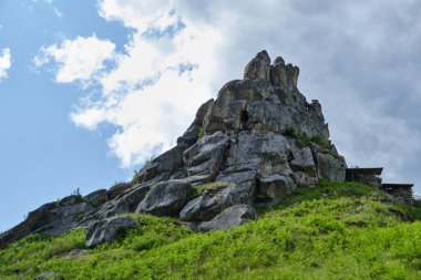Çimenli bir yamaçta yükselen engebeli kaya oluşumu. Parlak mavi gökyüzünün altında, dağılmış bulutlar, yaz güneşinde dağ manzarası..