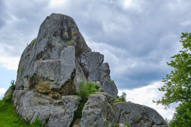 Çimenli bir yamaçta yükselen engebeli kaya oluşumu. Parlak mavi gökyüzünün altında, dağılmış bulutlar, yaz güneşinde dağ manzarası..